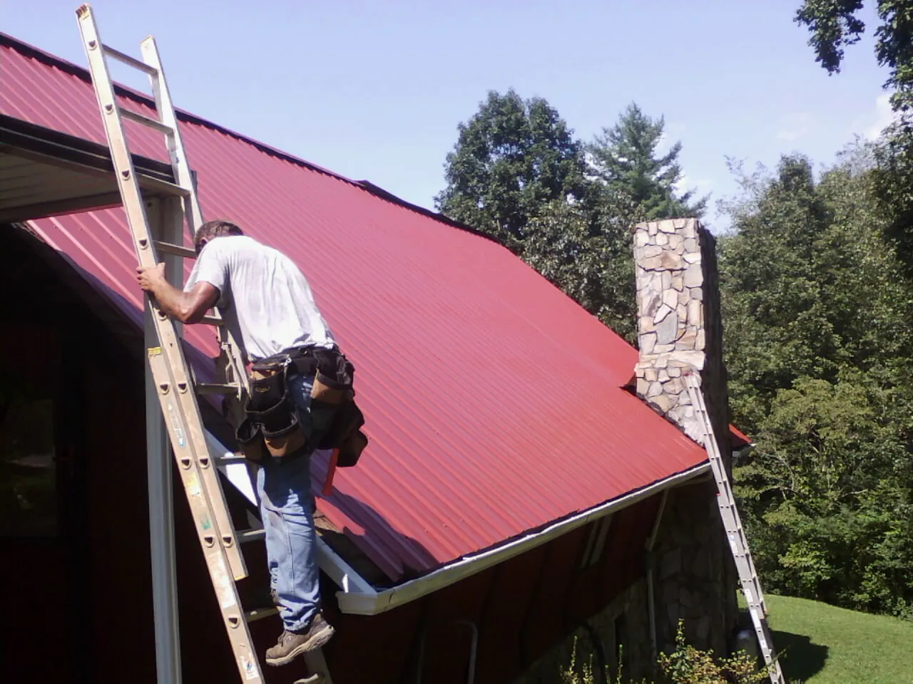 Roofer on a ladder beside a red metal roof and stone chimney — Soffit & Fascia Repair by Roofing Brothers in Glassport